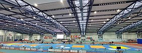 Eine große Sporthalle mit blauen und gelben Matten, die für Wettkämpfe vorbereitet sind. Aufbau von Tischen und Bildschirmen ist sichtbar. An den Wänden hängen verschiedene Flaggen. Die Decke hat eine moderne Stahlstruktur und ist gut beleuchtet.
