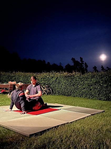 Zwei Männer sitzen auf einer roten Matte im Freien bei Nacht, umgeben von Gras und einer Hecke. Im Hintergrund leuchtet der Mond am Himmel. Die Szene strahlt eine entspannte und gesellige Atmosphäre aus.