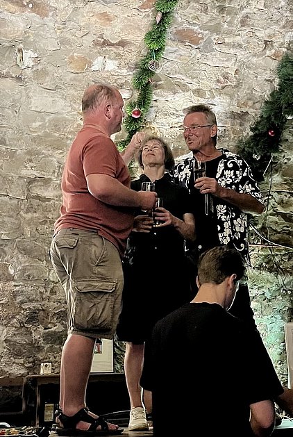 Drei Personen stehen in einem festlichen Raum, während sie Gläser anstoßen. Eine Frau in der Mitte hebt ihren Kopf, während zwei Männer, einer im T-Shirt und einer in einem karibischen Hemd, freundlich miteinander interagieren. Im Hintergrund sind Dekorationen und eine ungefähre Steinwand sichtbar.