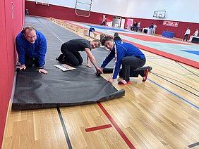 Drei Männer sind am Boden einer Sporthalle tätig. Sie rücken Matten zurecht; zwei knien dabei und ziehen die Matte, während einer mit einem Klebeband arbeitet. Im Hintergrund sind weitere Personen und Sportmatten sichtbar.