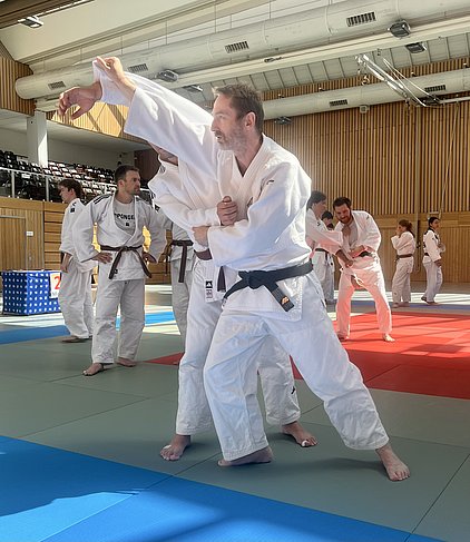 Eine Gruppe von Judokas in weißen Judogis übt in einem Trainingsraum. Im Vordergrund sind zwei Kämpfer zu sehen, wobei einer den anderen in einer Technik festhält. Die Atmosphäre ist konzentriert und aktiv, mit mehreren weiteren Judokas im Hintergrund, die ebenfalls trainieren.