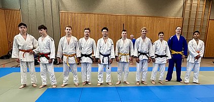 Eine Gruppe von zehn Judo-Kämpfern steht auf einer Matte, jeder in einer anderen Judo-Gi mit verschiedenen Gürtelfarben. Sie sind in zwei Reihen aufgestellt und blicken in die Kamera. Die Umgebung zeigt eine Judo-Halle mit Holzverkleidung im Hintergrund.