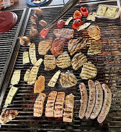Ein Grill mit verschiedenen Fleischsorten wie Steaks, Würstchen und Lachs, sowie gegrilltem Gemüse wie Zucchini und Paprika. Die Speisen sind goldbraun und mit Grillstreifen versehen. Rauch steigt auf, was auf das Grillen hinweist.