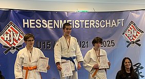 Drei Jungjudoka stehen stolz auf dem Siegespodest bei der Hessenmeisterschaft. Der erste Platz trägt eine goldene Medaille und hält eine Urkunde. Der zweite Platz hat eine orange und der dritte Platz eine blaue Gürtelfarbe. Im Hintergrund ist ein Banner mit der Aufschrift "Hessenmeisterschaft" zu sehen.