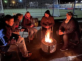 Eine Gruppe von fünf Personen sitzt in einem Kreis um ein Lagerfeuer. Sie sind in warme Kleidung gehüllt und genießen die Atmosphäre. Im Hintergrund sind verschwommene Konturen eines Sportfeldes bei Nacht zu sehen. Die Personen wirken entspannt und unterhalten sich.