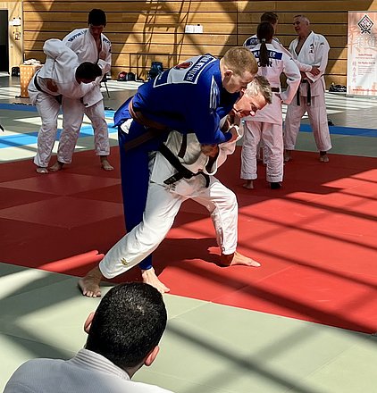 Eine Judo-Trainingsszene mit mehreren Kämpfern auf Matten. Im Vordergrund kämpft ein Athlet im blauen Gi mit einem anderen im weißen Gi. Im Hintergrund sind weitere Judokas beim Training zu sehen. Die Beleuchtung schafft eine dynamische Atmosphäre.