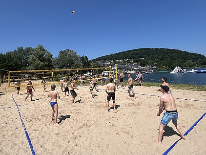 Eine Gruppe junger Männer spielt Beachvolleyball am Strand. Der Himmel ist klar und blau, das Spielfeld ist mit gelben Netzen und blauen Linien markiert. Im Hintergrund sind Bäume und Boote am Wasser zu sehen.