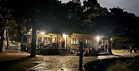 Eine hell erleuchtete Hütte steht im Dunkeln, umgeben von Bäumen. Gruppen von Menschen stehen auf der Veranda und interagieren miteinander. Der Weg zur Hütte ist gepflastert, und einige Fahrräder sowie andere Gegenstände sind sichtbar. Die Atmosphäre wirkt gesellig und einladend.