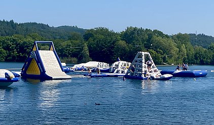 Eine Gruppe von Menschen spielt und schwimmt auf einer Vielzahl von aufblasbaren Wasserspielgeräten in einem See. Im Vordergrund sind eine Wasserrutsche und ein Kletterturm zu sehen, während andere schwimmende Geräte im Hintergrund angeordnet sind. Die Umgebung ist sonnig und grün.