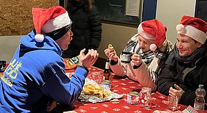 Drei Personen mit Weihnachtsmützen sitzen an einem Tisch, auf dem Essen und Getränke stehen. Eine Person isst etwas, während die anderen am Tisch miteinander sprechen und lachen. Die Atmosphäre wirkt festlich und gesellig.
