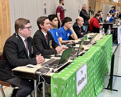 Eine Gruppe von fünf Personen sitzt an einem Tisch, der mit einem grünen Tuch bedeckt ist. Sie arbeiten an Laptops und scheinen Ergebnisse zu erfassen. Hinter ihnen steht eine weitere Gruppe von Personen. Auf dem Tisch steht ein Schild mit der Aufschrift „Matte 3“.