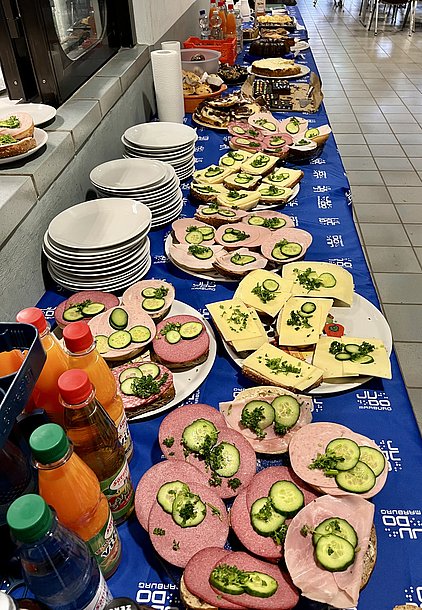 Ein Buffet mit verschiedenen belegten Broten, unter anderem mit Aufschnitt, Käse und Gurkenscheiben, ist auf einem Tisch angerichtet. Daneben stehen Getränke in bunten Flaschen. Die Stimmung vermittelt eine gesellige Essenszeit.