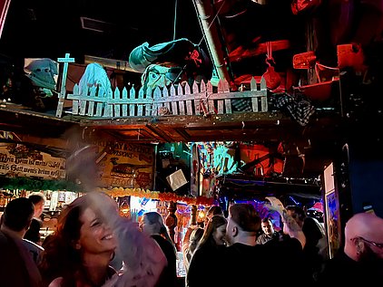 In einem lebhaften Club voller Menschen tanzen und lachen die Gäste. Der Raum ist mit bunten Lichtern dekoriert, und im Hintergrund sind kreative Dekorationen zu sehen, die eine festliche Atmosphäre schaffen. Ein fröhliches Ambiente, das Gemeinschaft und Spaß vermittelt.