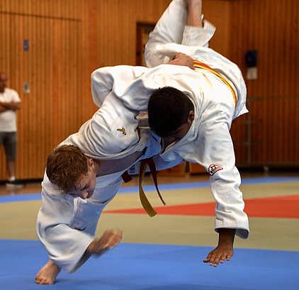 Zwei Judoka in weißen Anzügen führen einen Wurf aus. Der Athlet mit rotem Gürtel wird in die Luft geworfen, während der andere sich mit einer kraftvollen Bewegung abstützt. Die Szene findet auf einer Judomatte in einem Sportraum statt.