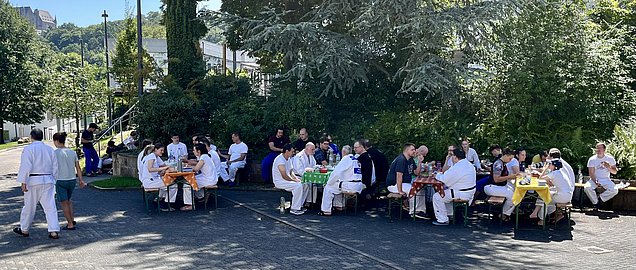 Eine Gruppe von Menschen in weißen Kleidung sitzt an mehreren Tischen im Freien. Sie genießen ein gemeinsames Essen und unterhalten sich. Die Szenerie ist von Bäumen und Sträuchern umgeben, im Hintergrund sind Gebäude sichtbar. Es herrscht eine fröhliche Atmosphäre.