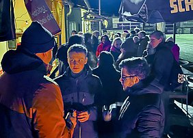 Eine Gruppe von Menschen steht in einer dunklen Umgebung, einige führen Gespräche. Im Vordergrund stehen zwei Männer, einer hält ein Mikrofon, während die andere Person aufmerksam zuhört. Im Hintergrund sind weitere Personen und Stände sichtbar, die eine gesellige Atmosphäre vermitteln.