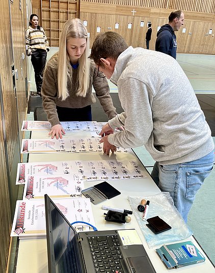Zwei Personen arbeiten an einem Tisch, auf dem Urkunden ausgelegt sind. Eine Frau mit langen, blonden Haaren betrachtet die Urkunden, während ein Mann in einem grauen Sweatshirt sie bearbeitet. Im Hintergrund sind weitere Personen sichtbar. Ein Laptop steht ebenfalls auf dem Tisch.