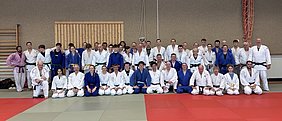 Eine Gruppe von Judokas in weißen und blauen Judogi steht in mehreren Reihen auf einer Matte. Die Teilnehmer sind verschiedene Altersgruppen und Geschlechter und posieren gemeinsam für ein Gruppenfoto in einer Sporthalle.