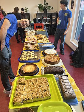 Ein Mensch mit einem freundlichen Lächeln steht neben einem Tisch voller Süßigkeiten, darunter verschiedene Kuchen, Kekse und Fantasietorten. Im Hintergrund sind weitere Personen und Pflanzen sichtbar, während die Umgebung eine einladende, gesellschaftliche Atmosphäre vermittelt.