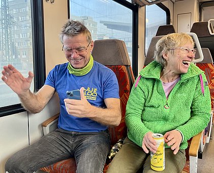 Zwei Personen sitzen in einem Zug und lachen fröhlich. Der Mann trägt ein blaues T-Shirt und eine grüne Halsschütze, während die Frau eine grüne Jacke anhat. Beide scheinen Spaß zu haben, während sie miteinander kommunizieren.