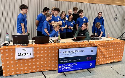 Eine Gruppe von Jugendlichen in blauen T-Shirts steht um einen Tisch mit zwei Laptops. An einem Laptop sitzt ein Trainer, der das Geschehen auf einem Bildschirm beobachtet, der die Namen und Punkte von zwei Judoka anzeigt. Der Wettkampf ist im Gange.
