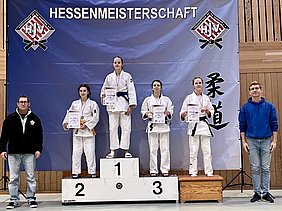 Das Bild zeigt eine Siegerehrung bei der Hessenmeisterschaft im Judo. Fünf Mädchen in Judogis stehen auf dem Podium. Die mittlere Teilnehmerin, mit einer blauen Gürtelfarbe, hält eine Medaille und eine Urkunde. Daneben stehen Trainer und Zuschauer.