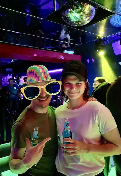 Zwei junge Männer stehen in einem beleuchteten Nachtclub. Der erste trägt eine bunte, große Sonnenbrille und einen gestreiften Hut. Beide halten Getränke in der Hand und lächeln in die Kamera. Im Hintergrund sind weitere Personen und schwache Lichter zu sehen.