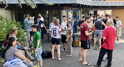 Eine Gruppe von Menschen steht und sitzt in einem Außenbereich. Einige halten Getränke in der Hand und unterhalten sich. Im Hintergrund sind Bänke und eine Gebäudefront sichtbar. Es scheint eine gesellige, entspannte Stimmung zu herrschen.