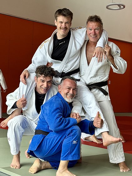 Vier Männer in BJJ-Gi posieren als Gruppe auf einer Judomatte. Zwei stehen hinten, während zwei vorne hocken. Alle lächeln und zeigen Daumen nach oben. Der Hintergrund ist hell rot.
