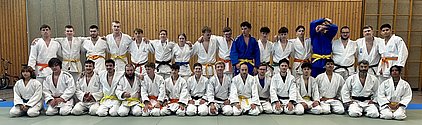 Eine Gruppe von 30 Judo-Athleten, hauptsächlich männlich, steht in weißer Judogewandung. Einige tragen farbige Gürtel (gelb, orange, blau, schwarz). Sie posieren gemeinsam auf einer Judo-Matte in einer Halle mit Holzverkleidung im Hintergrund.