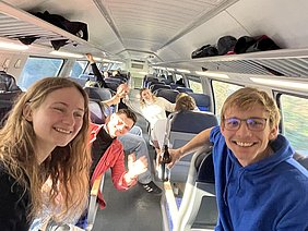 Eine Gruppe von Freunden sitzt in einem Zug und lächelt in die Kamera. Hinter ihnen sind weitere Passagiere zu sehen. Einige haben Getränke in der Hand und scheinen entspannt und fröhlich zu sein. Die Atmosphäre wirkt freundlich und gesellig.