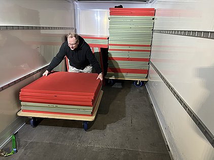 Ein Mann bringt Stapel aus roten und grünen Platten auf einem Wagen in einem Lkw. Im Hintergrund sind weitere gestapelte Platten zu sehen. Der Mann wirkt konzentriert, während er die Platten bewegt.