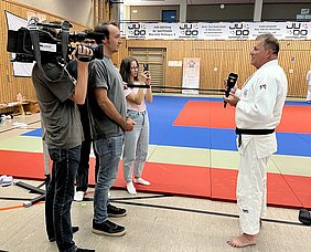 Ein Weißer-Gürtel Judo-Trainer spricht in einem Judo-Dojo mit einem Kamerateam und zwei weiteren Personen. Der Trainer hält ein Mikrofon und erklärt etwas, während die anderen aufmerksam zuhören. Der Boden ist mit bunter Judomatte ausgestattet, die verschiedene Farbbereiche aufweist.