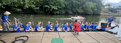 Eine Gruppe von Sportlern in blauen T-Shirts sitzt in einem Drachenboot an einem Gewässer. Einige Mitglieder halten Paddel, während andere lächeln. Im Hintergrund ist ein weiteres Boot zu sehen, das durch das Wasser fährt. Bäume umgeben die Szene.