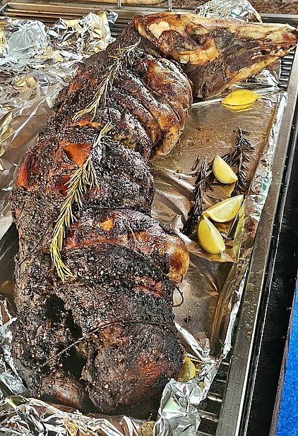 Ein auf einem Blech gegrilltes Stück Fleisch, das goldbraun und kross aussieht, ist mit Kräutern, darunter frischer Rosmarin, garniert. Neben dem Fleisch liegen einige Zitronenschnitze. Die Szene ist von glänzendem Alufolie-Hintergrund umgeben.