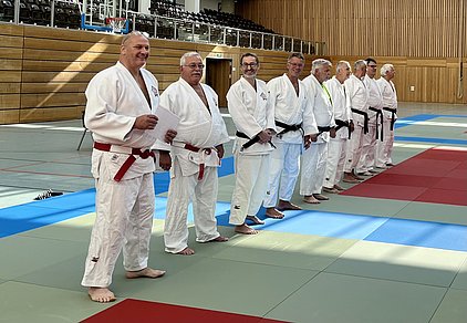 Eine Gruppe von acht Männern steht in einem Judogi auf einer Judomatte. Sie tragen verschiedene Gürtel, darunter rot und schwarz. Die Männer lächeln und scheinen auf eine Veranstaltung oder Zeremonie vorbereitet zu sein. Im Hintergrund ist eine Sporthalle sichtbar.