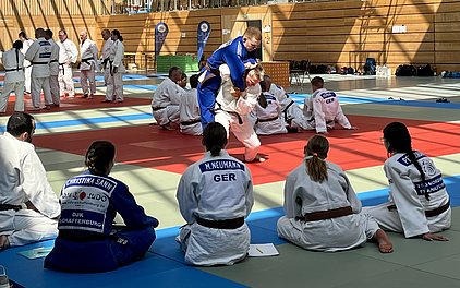 In der Turnhalle üben mehrere Judoka auf Matten. Ein Kämpfer im blauen Anzug führt einen Wurf aus, während andere in weißen Anzügen zuschauen. Der Fokus liegt auf der Technik und dem Training, um die Fähigkeiten im Judo zu verbessern.