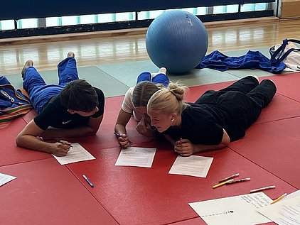 Drei Jugendliche liegen auf einer roten Matte und arbeiten gemeinsam an Aufgaben. Sie haben Stifte in der Hand und schreiben auf Blätter vor sich. Im Hintergrund ist ein Gymnastikball zu sehen.