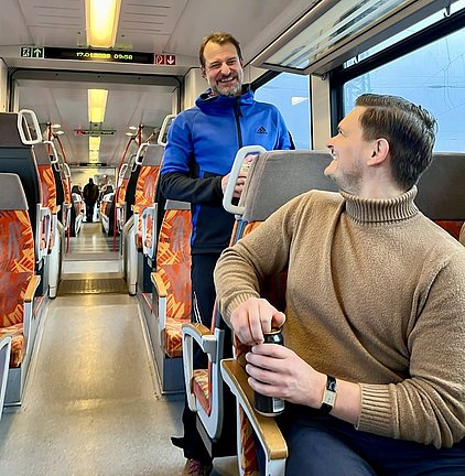 Zwei Männer in einem Zug sprechen miteinander und lächeln. Der Mann im blauen Oberteil steht, während der andere in einem beigen Pullover sitzt und eine Getränkedose in der Hand hält. Im Hintergrund sind leere Sitzplätze und ein weiteres Passagier zu sehen.