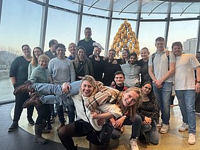 Eine Gruppe von 20 Menschen posiert in einem hellen Raum mit großen Fenstern und Blick auf einen Fluss. Einige Personen lächeln, während andere munter in die Kamera schauen. Im Hintergrund ist eine auffällige, goldene Skulptur zu sehen.