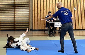 Zwei Kinder im Judoanzug kämpfen auf einer blauen Matte. Ein Trainer im blauen T-Shirt gibt Anweisungen und zeigt eine Handbewegung. Im Hintergrund sitzt ein Zeitnehmer am Tisch. Die Atmosphäre ist sportlich und konzentriert.