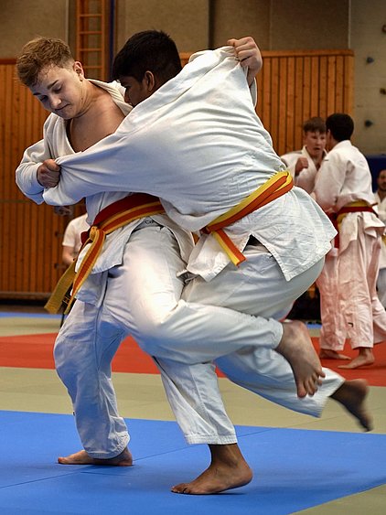 Zwei Judokas in weißen Anzügen kämpfen auf einer Matte. Einer hält den anderen fest, während beide auf den Füßen stehen. Im Hintergrund sind mehrere weitere Judokas zu sehen, die ebenfalls trainieren. Die Atmosphäre ist sportlich und dynamisch.